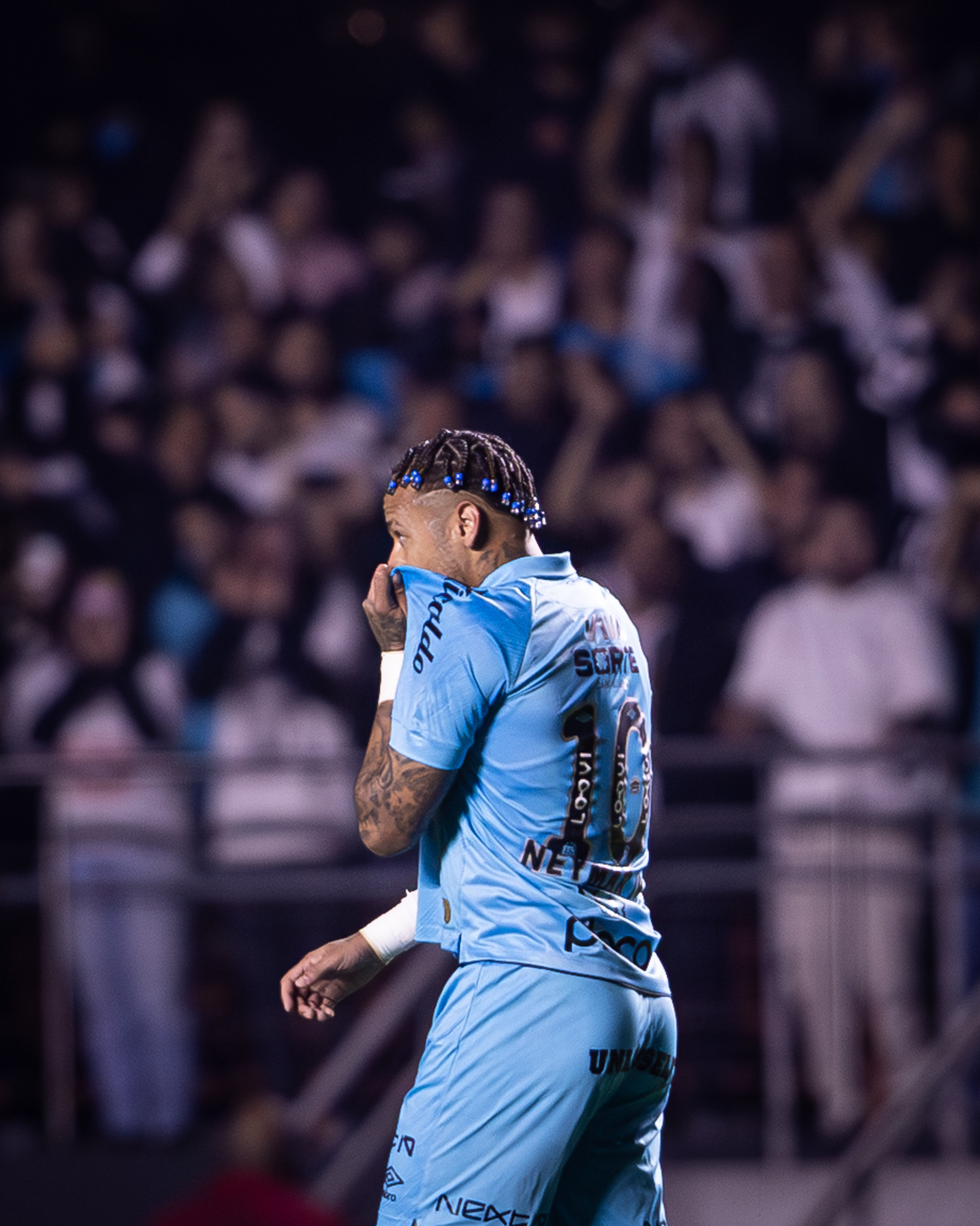 O futebolista brasileiro Neymar Jr., de costas e com a camisola azul número 10 do Santos F.C., cobre a boca e o nariz com a camisola enquanto está em campo num jogo noturno. Ele usa tranças com miçangas azuis. O estádio, cheio de adeptos desfocados, está no fundo.